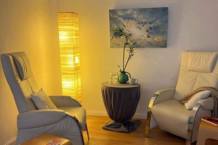 Auf dem Bild ist ein Beratungssetting zu sehen. Es besteht aus zwei Sesseln, die einander gegenüberstehen. zu jedem gibt es einen Beistelltisch. Im Hintergrund sieht man eine Stehlampe mit warmem Licht und ein Bild an der Wand.